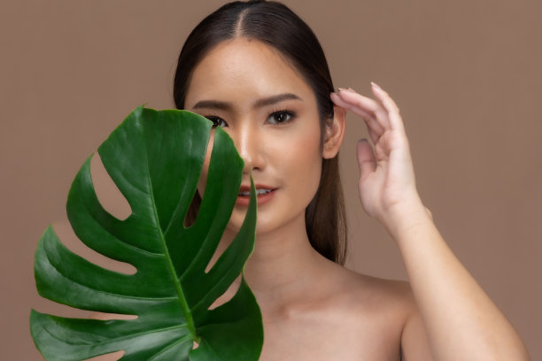 美丽的亚洲女人动人的眉毛微笑，干净和新鲜的皮肤在米色背景上幸福..美丽的肖像，迷人的年轻女孩与绿色的植物叶子隔离。图片下载