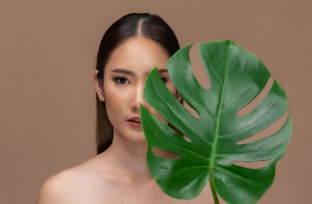 美丽的亚洲女人动人的眉毛微笑，干净和新鲜的皮肤在米色背景上幸福..美丽的肖像，迷人的年轻女孩与绿色的植物叶子隔离。图片下载