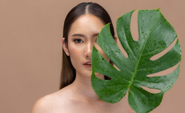 美丽的亚洲女人动人的眉毛微笑，干净和新鲜的皮肤在米色背景上幸福..美丽的肖像，迷人的年轻女孩与绿色的植物叶子隔离。图片下载