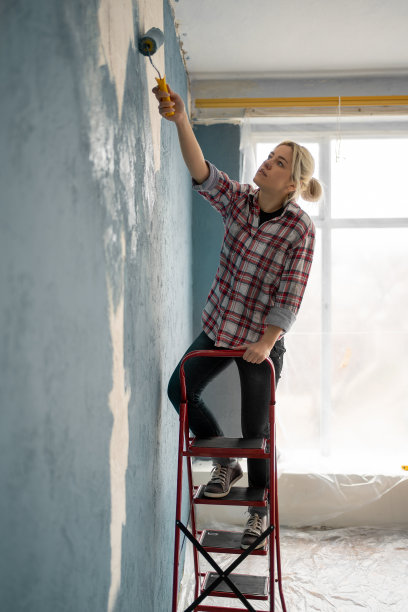 美丽的年轻女子在房间里画墙，站在梯子上的油漆辊。维修及房屋翻新图片下载