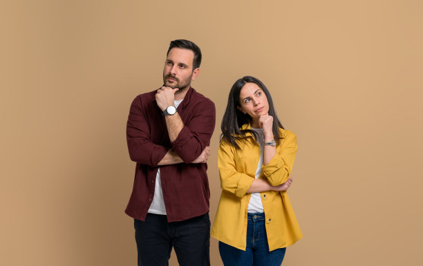 体贴的年轻夫妇穿着休闲装，抚摸着下巴，认真地摆姿势。困惑的男人和女人的肖像看着别处，而站在孤立的米色背景图片下载