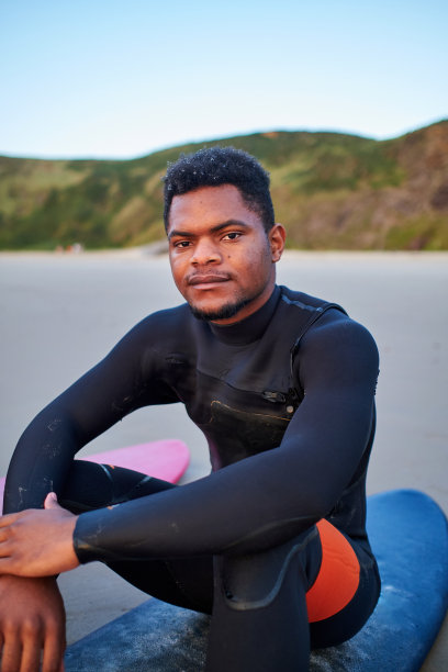 一个年轻的非洲裔美国人坐在沙滩上的冲浪板上看着相机的正面视图图片下载