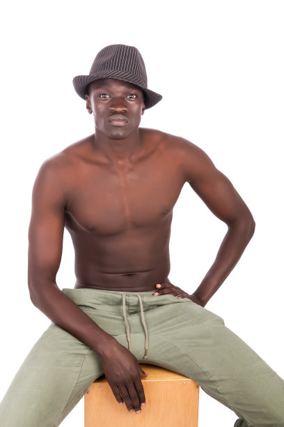Young buff Sudanese man with a naked torso in khaki trousers wearing a fedora sitting on a CajÃ³n drum.图片下载