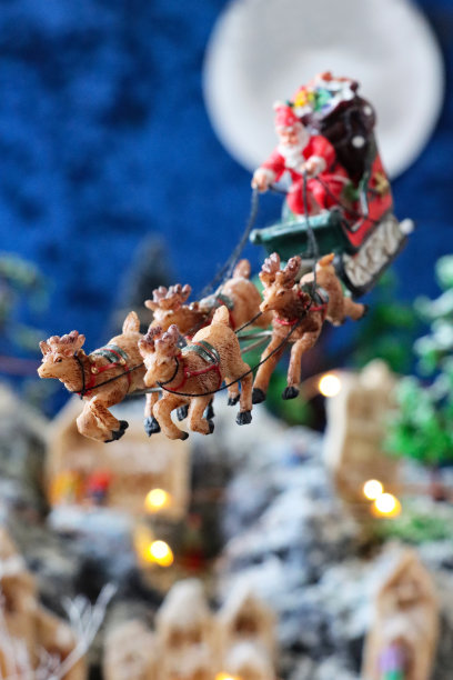 特写图像的圣诞老人雪橇和驯鹿飞越满月在风景优美的圣诞村显示，人工塑料和木制圣诞树在山上设置，照明自制木屋，重点在前景图片下载