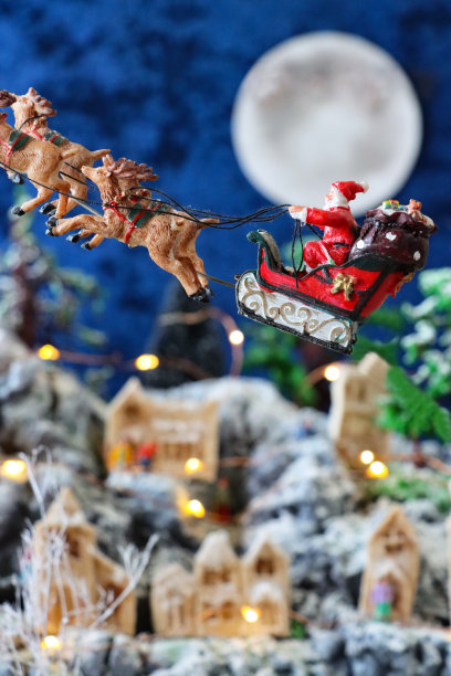 特写图像的圣诞老人雪橇和驯鹿飞越满月在风景优美的圣诞村显示，人工塑料和木制圣诞树在山上设置，照明自制木屋，重点在前景图片下载