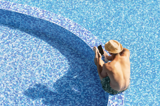 暑假期间，年轻人在度假酒店泳池边使用智能手机的俯视图。小伙拿着手机隔离在蓝水游泳池的背景复制空间。图片下载