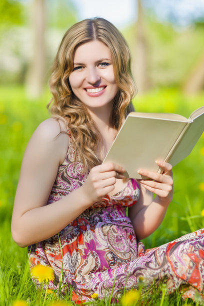 微笑的年轻迷人的白人金发女子阅读纸质书户外而坐在绿色的草地图片下载