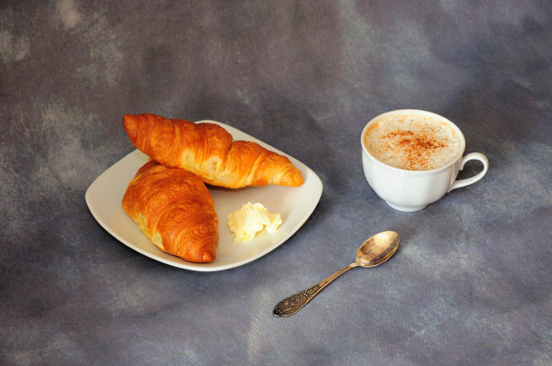 一杯加肉桂的热卡布奇诺，一个盘子里有两个新鲜的牛角包，灰色背景上有一块黄油。图片下载