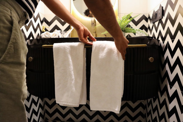 特写图像，一个无法辨认的人站在浴室的水槽边，把折叠的白色毛巾挂在连接到水槽梳妆台的毛巾架上，人造蕨类植物种在大理石浴室柜台上的金属花盆里，关注前景图片下载