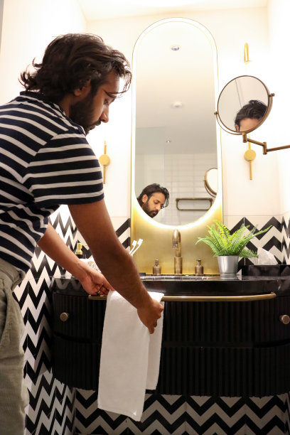特写图像，印度男子站在浴室的水槽上，将折叠的白色毛巾挂在毛巾栏杆上，连接到洗手台单元，镜子里的倒影，人造蕨类植物在大理石浴室柜台上的金属锅里，专注于前景图片下载
