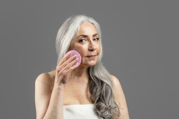 严肃的老白种人女士在毛巾清洁面部抬起海绵，孤立在灰色背景，工作室。美容护理图片下载