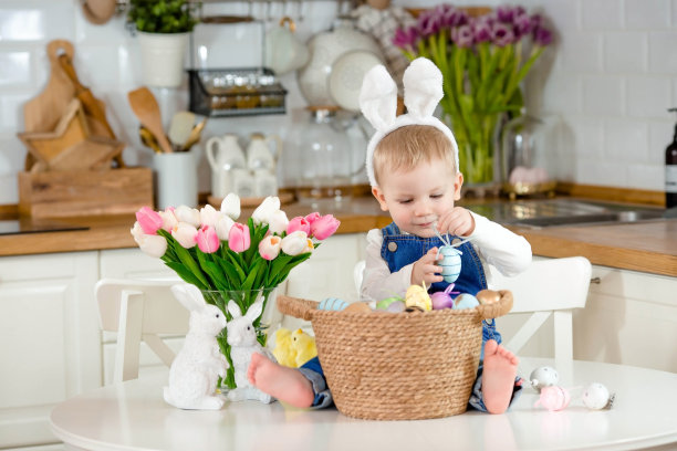 一个戴着兔子耳朵的小男孩，厨房桌子上放着一篮子复活节彩蛋。为家人准备复活节。孩子的笑脸。图片下载
