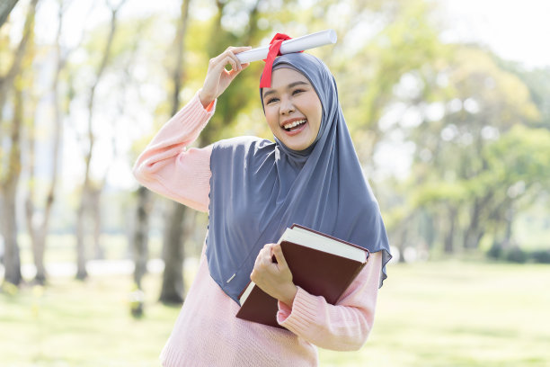 亚洲年轻的穆斯林女子戴着头巾庆祝毕业，手持书和学位卷，带着幸福的感觉，在公园微笑。祝贺教育理念图片下载