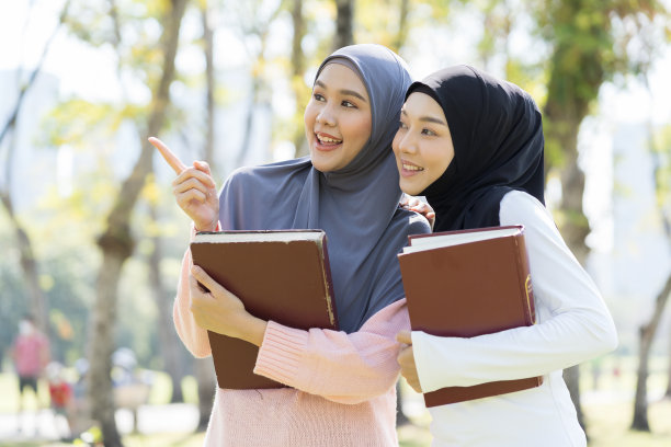两名年轻的亚洲穆斯林妇女戴着头巾，手拿着书，在公园里微笑。教育的概念图片下载