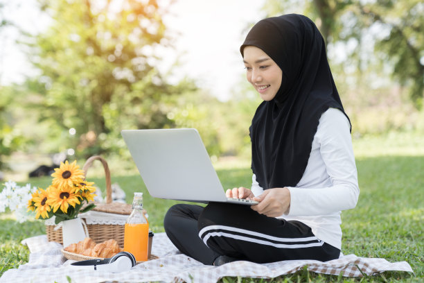 亚洲年轻的穆斯林妇女戴着头巾使用笔记本电脑与快乐和微笑坐在野餐公园在假日周末图片下载