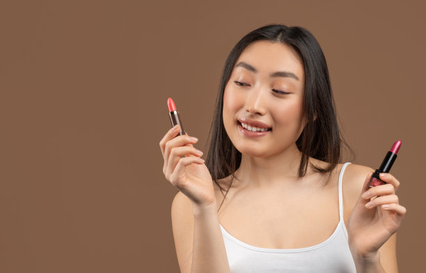 一名亚洲女子手里拿着两支口红，正在选择使用哪一支，在棕色的工作室背景下摆姿势图片下载
