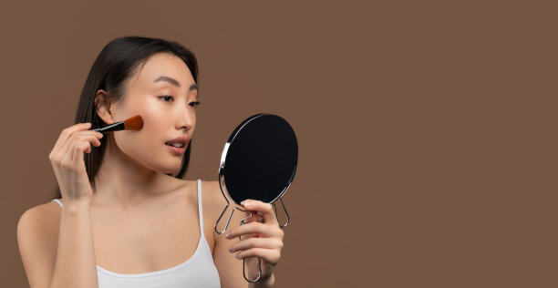 漂亮的亚洲女人用化妆刷在她的脸颊上涂腮红，看着镜子，棕色背景，全景图片下载