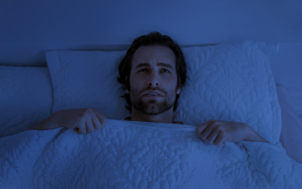 抑郁的年轻欧洲男子与胡茬躺在白色的床上毯子下的卧室内部在黑暗图片下载
