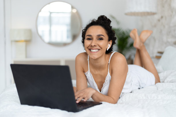 快乐的千禧一代白人棕发女士戴着无线耳机躺在柔软的床上，在电脑上打字图片下载