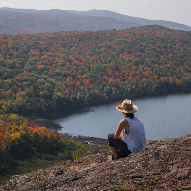 一位女性，戴着一顶草帽，穿着一件白衬衫，坐在岩石悬崖上，俯视着鲜艳多彩的秋天树木图片下载