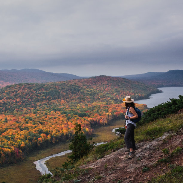 一位女徒步旅行者，戴着一顶草帽，站在一座岩石山上，俯瞰着明亮多彩的秋天的树木、河流和湖泊。密西根州了图片下载