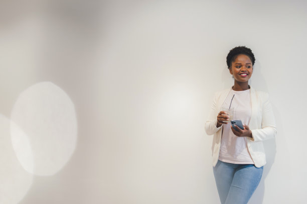 一名非裔美国妇女倚在有复印空间的白色墙壁上浏览社交媒体，喝冰咖啡。互联网信息。美味的早餐。互联网通信、无线连接技术。数字技术。图片下载