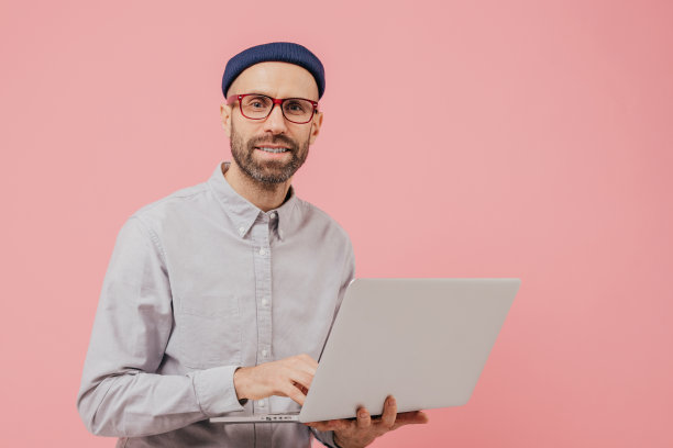 男子互联网用户在笔记本电脑上浏览网站的照片，连接到无线互联网，拿着电子产品，戴着眼镜，正式的衬衫，孤立在粉红色的背景与自由空间图片下载