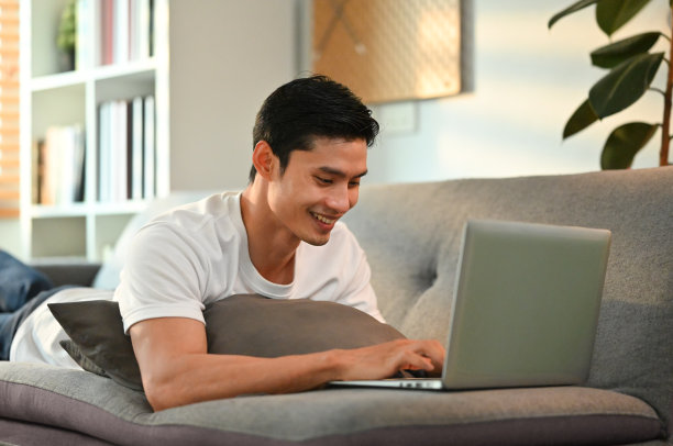 快乐的成年亚洲男人在休闲服在家工作，在笔记本电脑上浏览互联网，躺在客厅的沙发上。图片下载