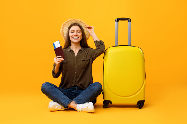 快乐的旅行者女子拿着护照坐在行李箱附近，黄色背景图片下载