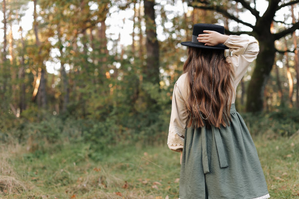 儿童女孩的后视图在时尚的帆布复古风格的连衣裙手握黑帽子在自然背景。小模特在公园里展示设计师的服装。照片目录。美与时尚理念图片下载