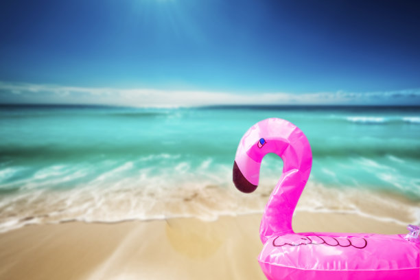 粉红色，时髦，吹海滩火烈鸟在赏金海滩与蓝色的海水背景。打夏天。粉红色的火烈鸟。粉红色火烈鸟，热带鸟类形状充气游泳池。图片下载