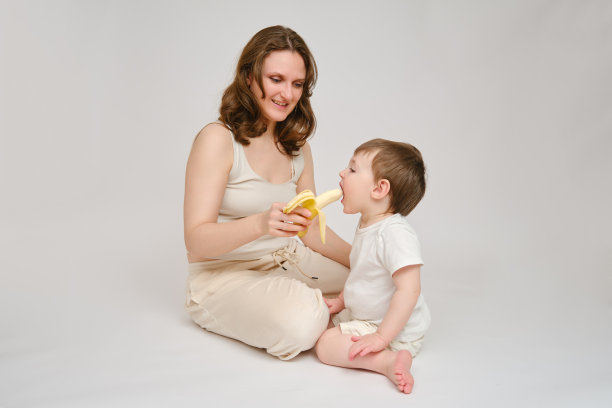 快乐的宝宝和妈妈吃香蕉在工作室白色背景。一个微笑的孩子和妈妈吃水果的照片。2岁左右(1岁9个月)图片下载