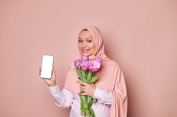 戴着粉色头巾的穆斯林妇女，手里拿着一部带有白色空白数字屏幕的智能手机，上面有移动应用程序的复制空间图片下载