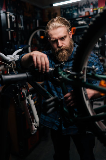 在室内黑暗的自行车修理店里，大胡子的野蛮修理工站在自行车架上修理山地车。图片下载