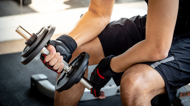 在健身房里，穿着黑色运动服，戴着运动手套，坐在锻炼板凳上举哑铃的运动男子。重量训练和健美概念。图片下载