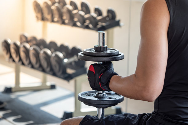 在健身房里，穿着黑色运动服，戴着运动手套，坐在锻炼板凳上举哑铃的运动男子。重量训练和健美概念。图片下载