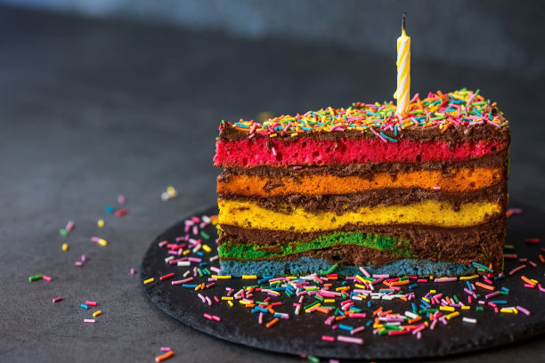 用节日蜡烛和五彩纸屑装饰的彩虹生日蛋糕。糖果。五颜六色的甜点，在黑色的背景上彩虹的颜色。图片下载