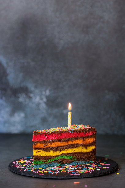 用节日蜡烛和五彩纸屑装饰的彩虹生日蛋糕。糖果。五颜六色的甜点，在黑色的背景上彩虹的颜色。图片下载