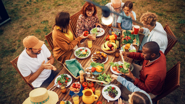 家人和朋友在家外庆祝的俯视高架景观。一群不同的孩子，成年人和老年人坐在一张桌子上，有有趣的对话。吃烧烤和蔬菜。图片下载