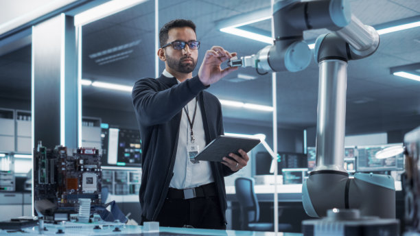 一个年轻的多民族男性工程师使用平板电脑的肖像。与机械臂机器交互，接收来自手部的微芯片。高科技工业实验室设施。图片下载