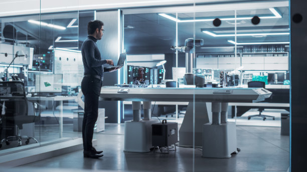 在一家高科技工厂里，工业机器人工程师站在机械臂旁边。男科学家用笔记本电脑操纵和编程机器人拿起并移动一个微芯片。图片下载