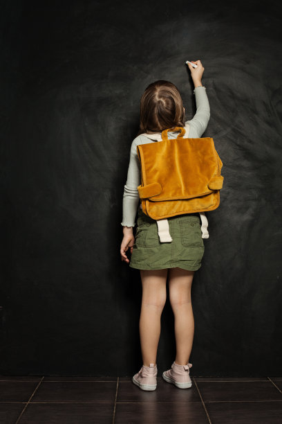背着橙色双肩包的小女孩站在那里，在黑色的学校黑板上写字。回到学校，接受教育，学生，学生图片下载