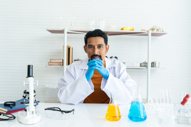 一位男老师穿着白色的实验大褂，戴着橡胶手套，架子和桌子上放着许多实验工具。桌子上有蓝色和黄色的化学烧瓶。双手合拢，微笑，直视前方。图片下载