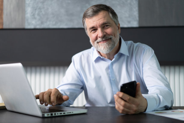 微笑的中年商人坐在办公室里拿着智能手机。中年经理CEO使用手机移动应用和笔记本电脑。用于业务发展的数字技术应用和解决方案图片下载