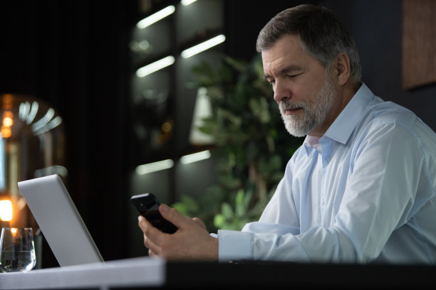 微笑的中年商人坐在办公室里拿着智能手机。中年经理CEO使用手机移动应用和笔记本电脑。用于业务发展的数字技术应用和解决方案图片下载
