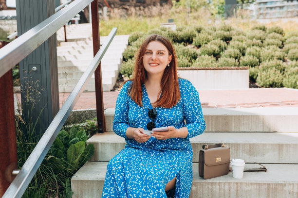 非常快乐的红发女人站在城市背景上，手里拿着车票和手机。女人手里拿着公交或地铁车票，女孩坐在城市街道的楼梯上。图片下载