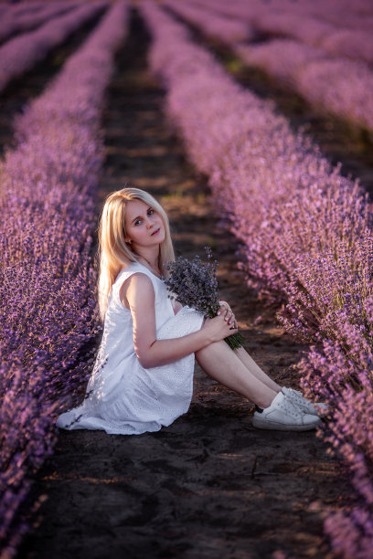 身穿白色连衣裙的年轻女子坐在田野里一排排紫色的薰衣草丛中。捧着花束的千禧女孩图片下载