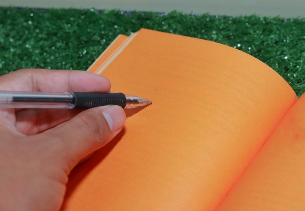 那只手握着笔，准备在一本橘色的空本子上写字图片下载