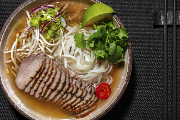 亚洲拉面汤，有小牛肉、香菜、面条、辣椒、切碎的葱、小绿芽和青柠角。用圆形、深棕色的陶瓷碗盛汤。碗立在一个黑色的柳条架子上。图片下载