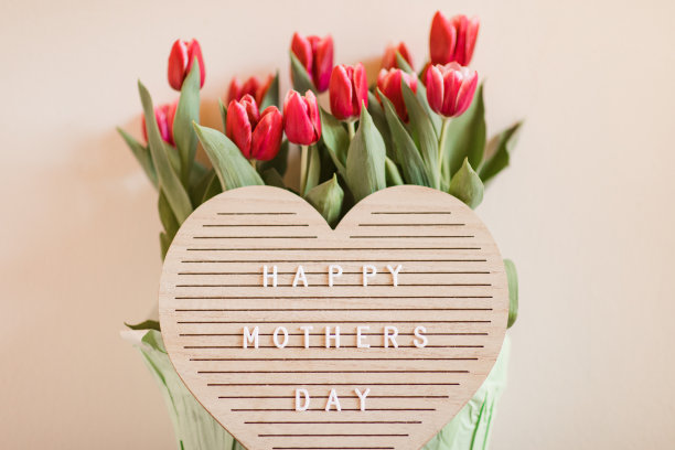 一个心形的字母板，上面写着“母亲节快乐”，明亮的粉红色郁金香盛开，郁郁葱葱的绿色茎和叶子在它后面，在明亮的自然日光下拍摄，中性的米色背景与复制空间图片下载